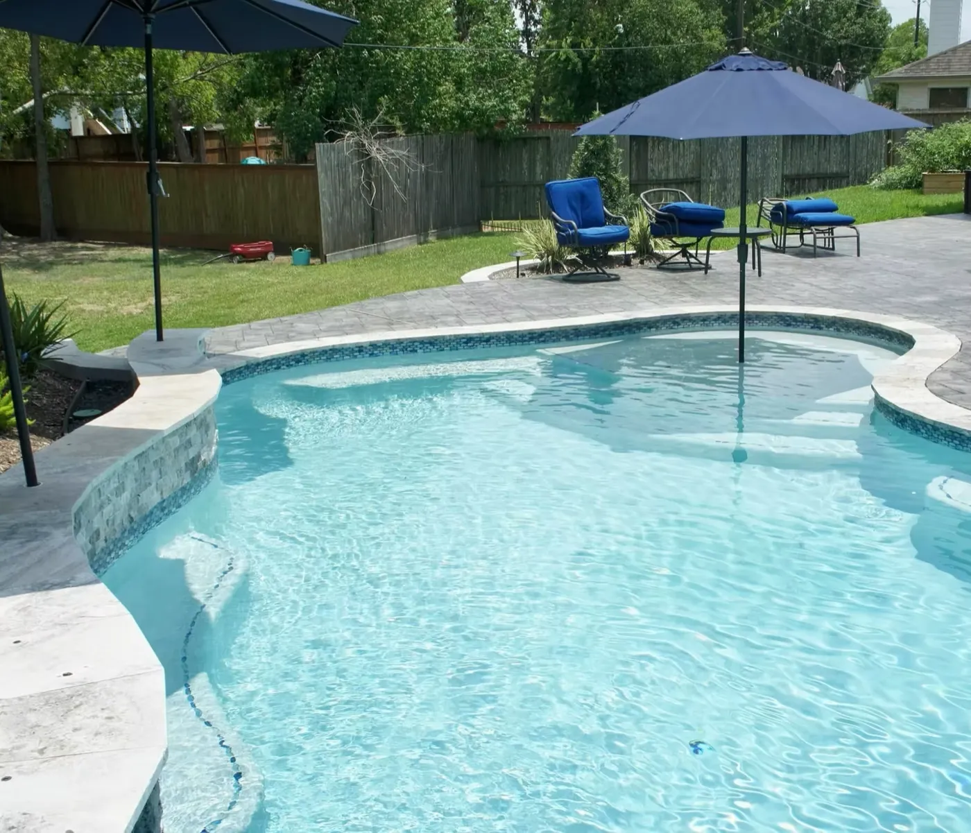 Custom freeform pool with blue umbrellas and stamped concrete patio built by Aquatic Pools in League City TX